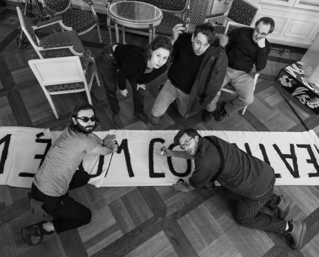 Czarno-białe zdjęcie. Kilka osób zgromadzonych wokół rozłożonego na podłodze długiego transparentu. Dwóch mężczyzn klęczy na parkiecie i domalowuje czarne litery na białym materiale, tworząc hasło „Teatr mój widzę, wolny”. Obok nich stoją i siedzą inne osoby, obserwując pracę. W tle widać stylowe krzesła i stolik.